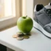 Stapel euromunten naast een groene appel en hardloopschoen op wit bureau, symboliek voor gezondheid en financiën.