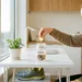 Glimlachende medewerker die gouden munt in glazen pot met munten dropt op modern bureau, groene plant en sportschoenen op de achtergrond.