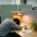 Uitgeputte werknemer hangend over modern bureau met halfleeg koffiekopje, onaangeroerde welzijnsbrochure en verwelkte plant in warm lamplich