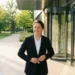Werknemer stapt ontspannen naar buiten bij modern kantoorgebouw, jas losjes open, groen park en gouden zonlicht op de achtergrond.
