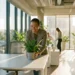 Medewerker plaatst groene plant op wit bureau in modern kantoor met warm ochtendlicht door grote ramen.