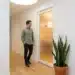 Medewerker in casual kantoorkleding stopt in de gang bij een zachtgloeiend wellnessdisplay aan een witte muur met groene plant.